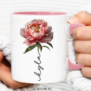 May include: White ceramic mug with a pink interior and handle. The mug features a pink peony flower with green leaves and the word "Kayla" written in cursive on the front. The mug is 11 oz.