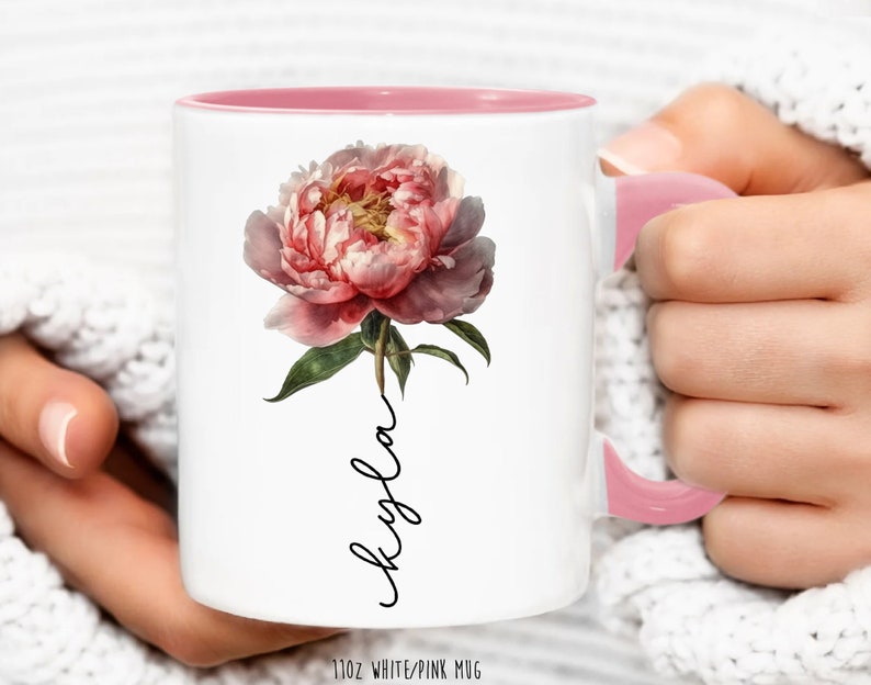 May include: White ceramic mug with a pink interior and handle. The mug features a pink peony flower with green leaves and the word "Kayla" written in cursive on the front. The mug is 11 oz.
