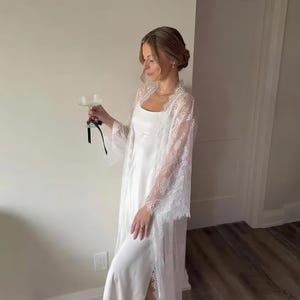 Könnte beinhalten: Ein weißes Satin-Slip-Kleid mit passendem Spitzen-Robe. Die Robe hat weite Ärmel und aufwendige florale Spitzendetails. Die Frau hält ein Champagnerglas mit einem schwarzen Riemen.