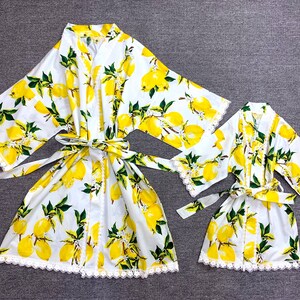 May include: Two white robes featuring a vibrant yellow lemon and green leaf pattern. Both garments include wide sleeves, a matching tie belt at the waist, and delicate white lace trim along the front edges, sleeve cuffs, and the bottom hemline.