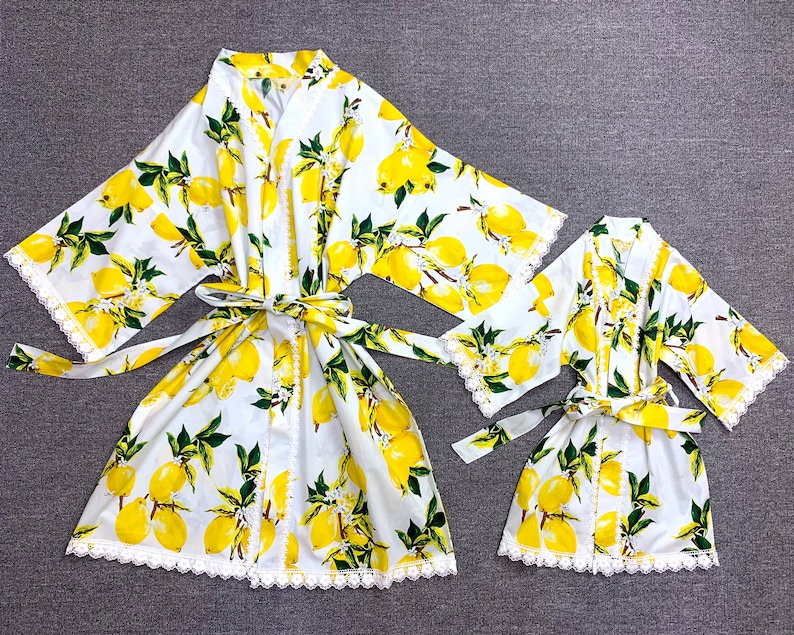 May include: Two white robes featuring a vibrant yellow lemon and green leaf pattern. Both garments include wide sleeves, a matching tie belt at the waist, and delicate white lace trim along the front edges, sleeve cuffs, and the bottom hemline.