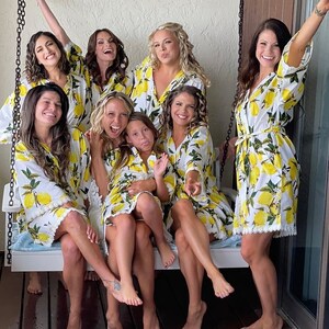 May include: A group of eight individuals wearing matching white robes featuring a vibrant yellow lemon and green leaf print. The robes have three-quarter length sleeves, a tie-waist closure, and a delicate white lace trim along the bottom hem.