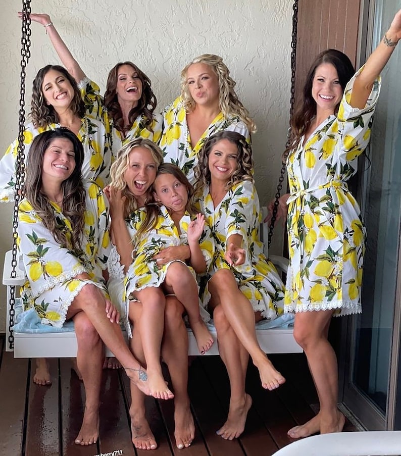 May include: A group of eight individuals wearing matching white robes featuring a vibrant yellow lemon and green leaf print. The robes have three-quarter length sleeves, a tie-waist closure, and a delicate white lace trim along the bottom hem.