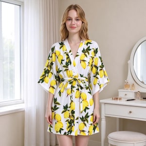 May include: A white short robe featuring a vibrant all-over print of yellow lemons and green leaves. The garment has wide three-quarter length sleeves, a V-neckline, and a matching fabric tie belt that secures at the waist.