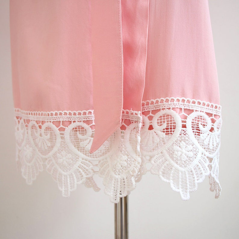 May include: A light pink robe featuring a smooth fabric texture and a matching tie belt. The hem is finished with a wide, intricate white lace trim that displays a detailed scalloped pattern with floral and scrollwork motifs throughout.