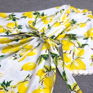 May include: A white robe featuring a vibrant yellow lemon and green leaf print with white floral accents. The garment includes a matching fabric tie belt at the waist and delicate white lace trim along the sleeve cuffs and front opening edges.