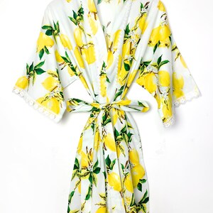 May include: A white robe featuring a vibrant yellow lemon and green leaf print pattern. The garment includes wide sleeves, a matching fabric waist tie, and delicate white lace trim along the front edges, sleeve cuffs, and the bottom hemline.
