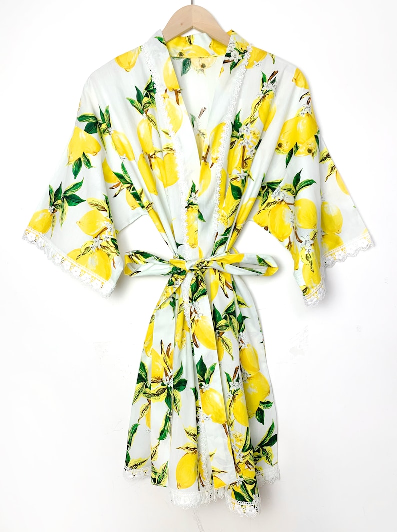 May include: A white robe featuring a vibrant yellow lemon and green leaf print pattern. The garment includes wide sleeves, a matching fabric waist tie, and delicate white lace trim along the front edges, sleeve cuffs, and the bottom hemline.