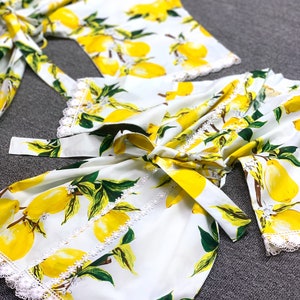 May include: A white robe featuring a vibrant yellow lemon and green leaf print pattern. The garment includes a matching fabric tie belt at the waist and delicate white lace trim detailing along the sleeve cuffs and the front opening edges.