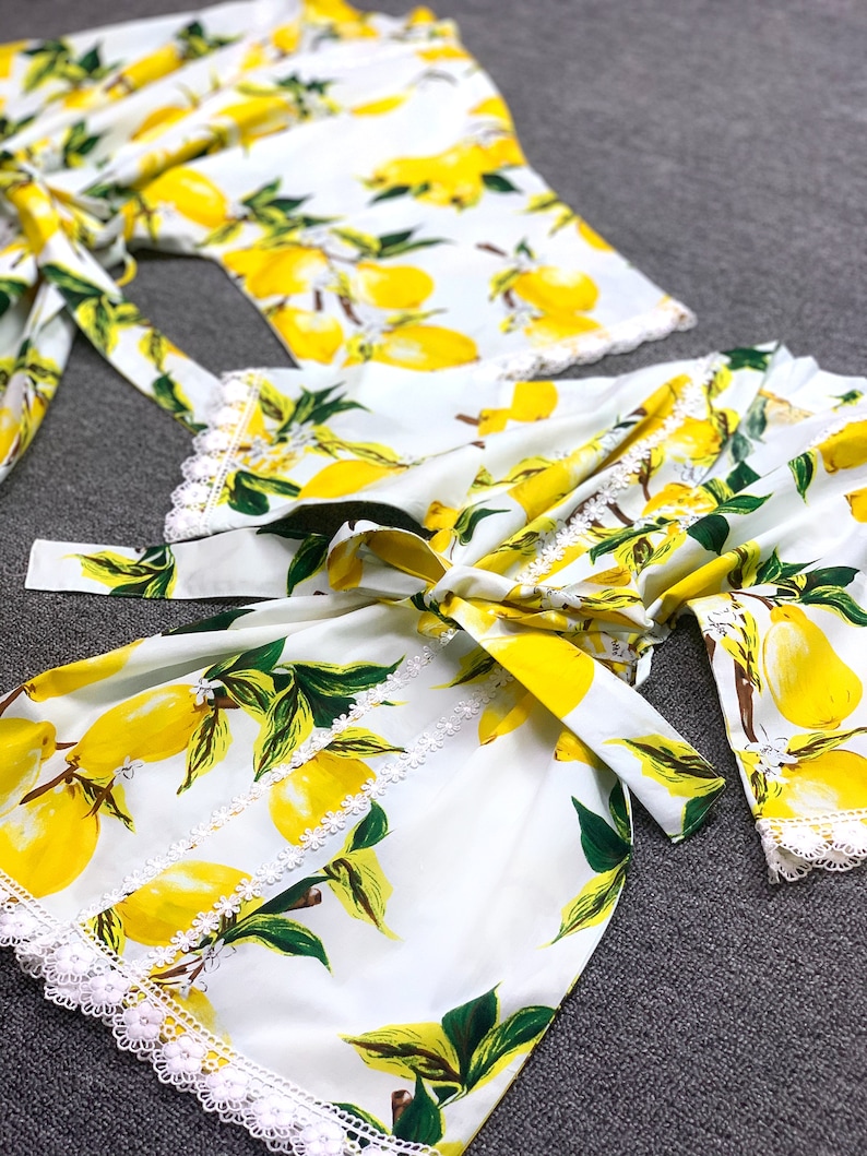 May include: A white robe featuring a vibrant yellow lemon and green leaf print pattern. The garment includes a matching fabric tie belt at the waist and delicate white lace trim detailing along the sleeve cuffs and the front opening edges.