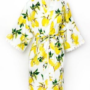 May include: A white robe featuring a vibrant yellow lemon and green leaf print pattern. The garment includes wide sleeves, a matching tie belt at the waist, and delicate white lace trim detailing along the sleeve cuffs and the front opening edges.