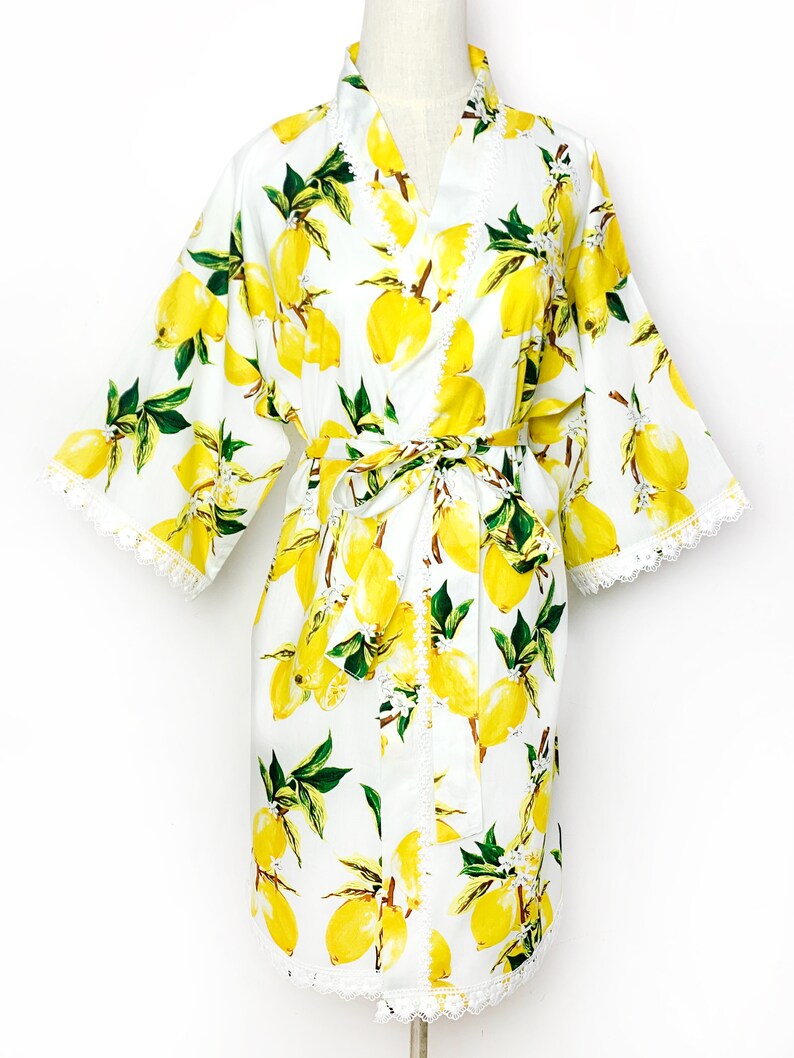 May include: A white robe featuring a vibrant yellow lemon and green leaf print pattern. The garment includes wide sleeves, a matching tie belt at the waist, and delicate white lace trim detailing along the sleeve cuffs and the front opening edges.