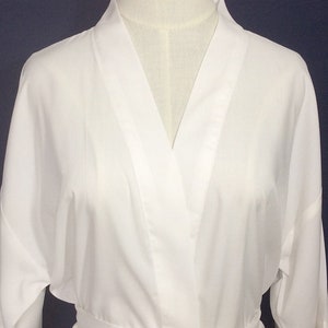 May include: A solid white robe featuring a classic wrap-style front with a wide collar and long sleeves. The garment is displayed on a mannequin, showcasing a smooth, clean fabric texture and a simple, structured design with a tie waist closure.