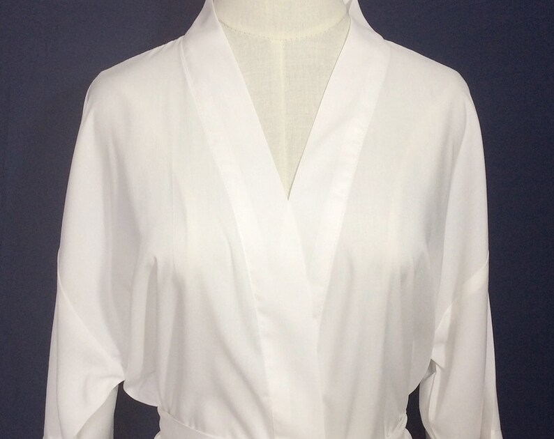 May include: A solid white robe featuring a classic wrap-style front with a wide collar and long sleeves. The garment is displayed on a mannequin, showcasing a smooth, clean fabric texture and a simple, structured design with a tie waist closure.