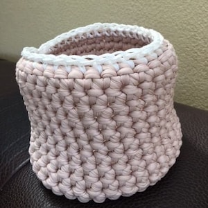 Könnte beinhalten: Ein rosa gehäkelter Korb mit einem weißen gehäkelten Rand. Der Korb ist aus dickem Garn gefertigt und hat eine runde Form.