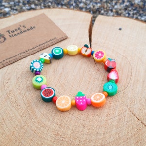 Peut inclure: Un bracelet coloré sur le thème des fruits avec des perles en forme de pastèque, de kiwi, de fraises, de citrons et d'oranges. Le bracelet est composé de perles colorées et est enfilé sur un cordon élastique transparent.