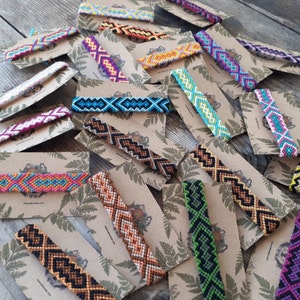 May include: Assortment of colorful friendship bracelets with geometric patterns, displayed on brown card stock with green leaf designs.