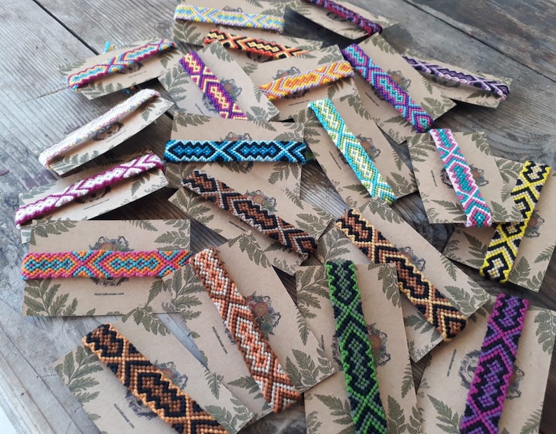 May include: Assortment of colorful friendship bracelets with geometric patterns, displayed on brown card stock with green leaf designs.