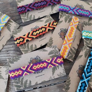 May include: Assortment of colorful friendship bracelets with geometric patterns, displayed on brown card stock with a leafy design.