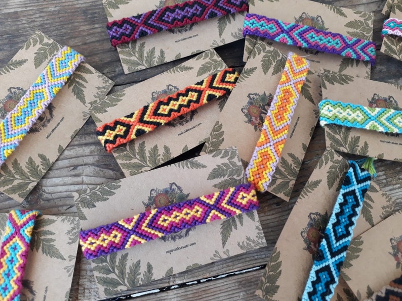 May include: Assortment of colorful friendship bracelets with geometric patterns, displayed on brown card stock with a leafy design.