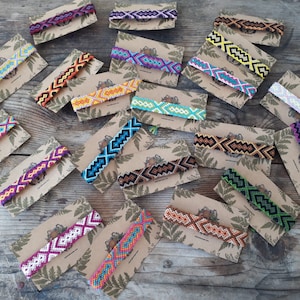 May include: A collection of colorful friendship bracelets with geometric patterns, displayed on brown card stock with a fern leaf design. The bracelets are made with colorful embroidery floss and feature a variety of colors, including red, orange, yellow, green, blue, purple, and black.