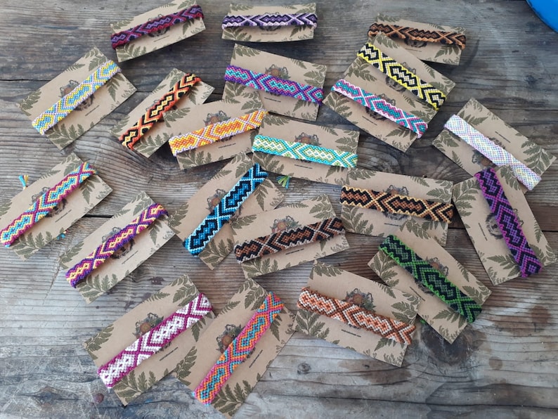 May include: A collection of colorful friendship bracelets with geometric patterns, displayed on brown card stock with a fern leaf design. The bracelets are made with colorful embroidery floss and feature a variety of colors, including red, orange, yellow, green, blue, purple, and black.