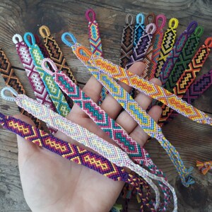 May include: A collection of colorful friendship bracelets with geometric patterns. The bracelets are made with colorful embroidery floss and have a variety of colors, including red, orange, yellow, green, blue, purple, pink, and white.