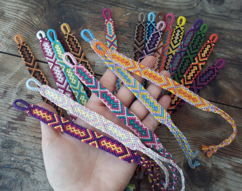 May include: A collection of colorful friendship bracelets with geometric patterns. The bracelets are made with colorful embroidery floss and have a variety of colors, including red, orange, yellow, green, blue, purple, pink, and white.