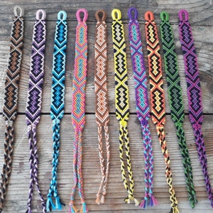 May include: A set of ten colorful friendship bracelets with various geometric patterns. The bracelets are made with colorful embroidery floss and have a knotted fringe at the ends.