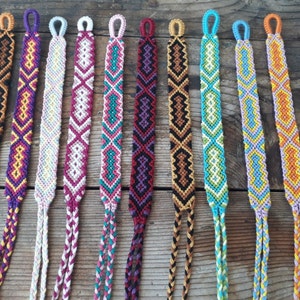 May include: Ten colorful friendship bracelets with different patterns and colors, including purple, pink, white, teal, brown, orange, and yellow.