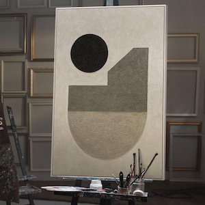 Puede incluir: Una pintura abstracta sobre un caballete en un estudio de arte. La obra presenta un círculo negro y formas geométricas en tonos grises y beige. Pinceles y una paleta están sobre una pequeña mesa. El estudio tiene varios marcos de fotos vacíos en la pared.
