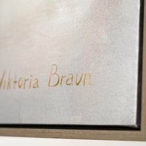 May include: Close-up of a white canvas with a gold signature that reads "Viktoria Braun". The canvas is framed in a light brown wood frame.