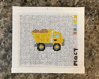 May include: A square needlepoint canvas featuring a yellow dump truck with a brown load, grey wheels, and a light blue window. A vertical color palette of yellow, light blue, grey, and brown squares is on the right, with the word "macy" printed below.