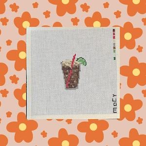 May include: A square piece of white needlepoint canvas featuring a stitched design of a brown iced beverage in a glass. The drink includes a red straw and a green lime wedge garnish. The canvas has a grid pattern and a small color palette on the side.