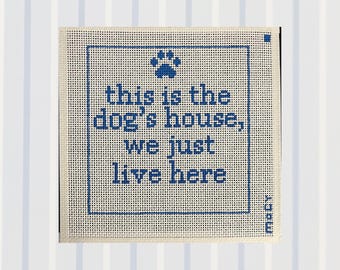 May include: A square needlepoint canvas featuring a blue paw print icon and the text "this is the dog's house, we just live here" in a blue block font. The design is enclosed within a blue rectangular border on a cream-colored mesh fabric.