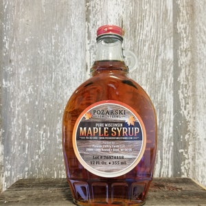 May include: A glass bottle of amber-colored maple syrup with a red lid. The label reads "Pozarski Family Farms Pure Wisconsin Maple Syrup" with the company's website and contact information. The bottle contains 12 fluid ounces or 355 milliliters of syrup.