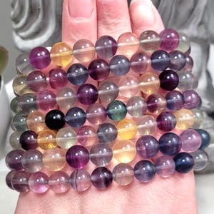 May include: A stack of rainbow fluorite gemstone bracelets. The bracelets are made with round beads and have a variety of colors, including purple, green, yellow, and clear.