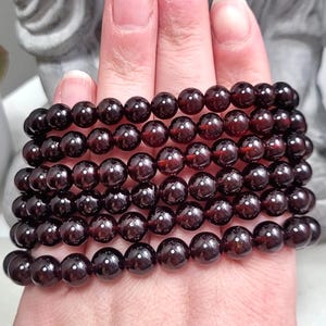 May include: A stack of eight garnet gemstone bracelets, each with round beads, on a person's hand. The bracelets are a deep red color.