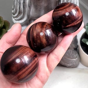 May include: Three red tiger's eye spheres, each with a different pattern of brown and gold stripes. The spheres are smooth and polished, and they are arranged in a row on a hand.