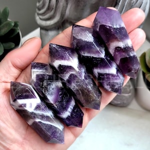 May include: A collection of six amethyst crystal wands. The wands are a deep purple with white and clear bands. The crystals are held in a hand, with a blurred background of plants and a statue.