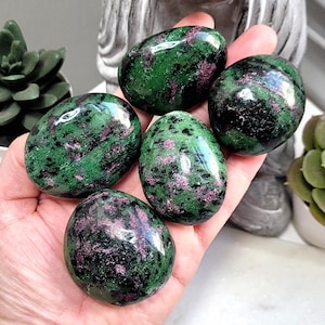 May include: Five polished ruby in zoisite stones, each with a unique pattern of green and pink hues. The stones are smooth and egg-shaped, held in an open hand. The background includes a succulent plant and a statue.