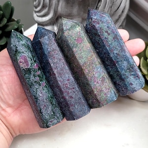 May include: Four polished ruby zoisite crystal towers. The crystals are a mix of dark green, blue, and grey, with flecks of red ruby. Each tower is approximately 10 cm long and has a pointed top. The crystals are held in a hand.