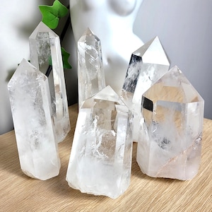 May include: A collection of clear quartz crystal points of varying sizes. The crystals are faceted and have a polished appearance. They are arranged on a light-colored wooden surface, with a white bust and green leaves in the background.