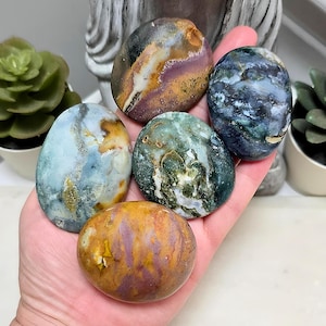 Peut inclure: Cinq pierres polies, lisses et de forme ovale, présentant des motifs et des couleurs naturels variés, notamment des nuances de violet, jaune, bleu, vert et blanc, reposant dans une paume. Chaque pierre présente des inclusions minérales uniques.