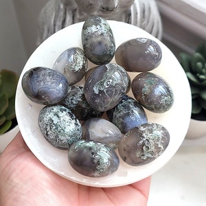 May include: A collection of moss agate tumbled stones in various shades of green and grey. The stones are smooth and polished, with a natural, organic look.