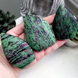 May include: Three polished zoisite stones with a green base colour and black and pink inclusions. The stones are teardrop-shaped and held in a hand. A grey Buddha statue and a succulent plant are in the background.