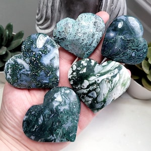 Peut inclure: Une main tient six pierres en forme de cœur. Les pierres sont principalement vertes avec des veines blanches et bleues, ressemblant à de l'agate mousse. Une statue de Bouddha grise et des plantes succulentes en pot sont en arrière-plan.