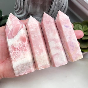 Peut inclure: Quatre tours en opale rose polie avec une pointe hexagonale. Chaque pierre présente des variations naturelles de couleur, allant du rose pastel doux au blanc crème, avec des surfaces lisses et réfléchissantes et des textures cristallines.