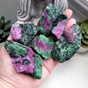 May include: A hand holds several rough-cut ruby in zoisite stones. The stones display a mix of deep green and black hues, with vibrant patches of pink and magenta. The gemstones are natural and unpolished.