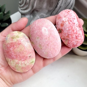 May include: Three oval stones in pink and red hues are shown. The stones have a smooth, polished finish with natural colour and texture variations. They are held in a hand, with a blurred background of greenery and a statue.